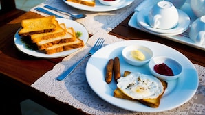 Desayuno con tostadas, huevos, salchichas y mermeladas en platos, junto con una tetera y tazas sobre una mesa.