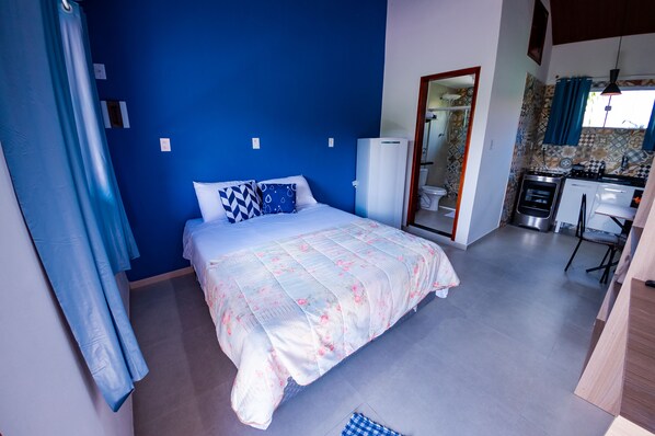 A bedroom with a bed, a blue wall, a small kitchenette, and a bathroom visible through an open door.