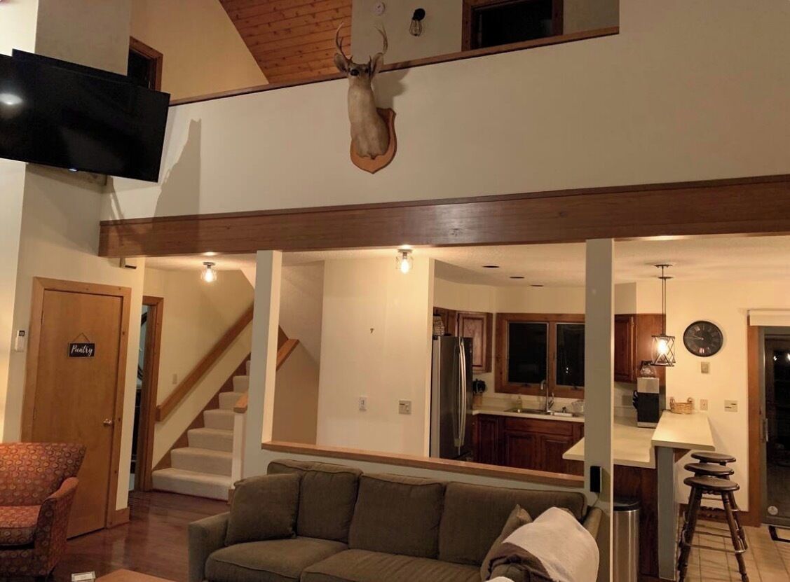 A living room with a sofa, a mounted deer head, a TV, and a staircase leading to a kitchen area.