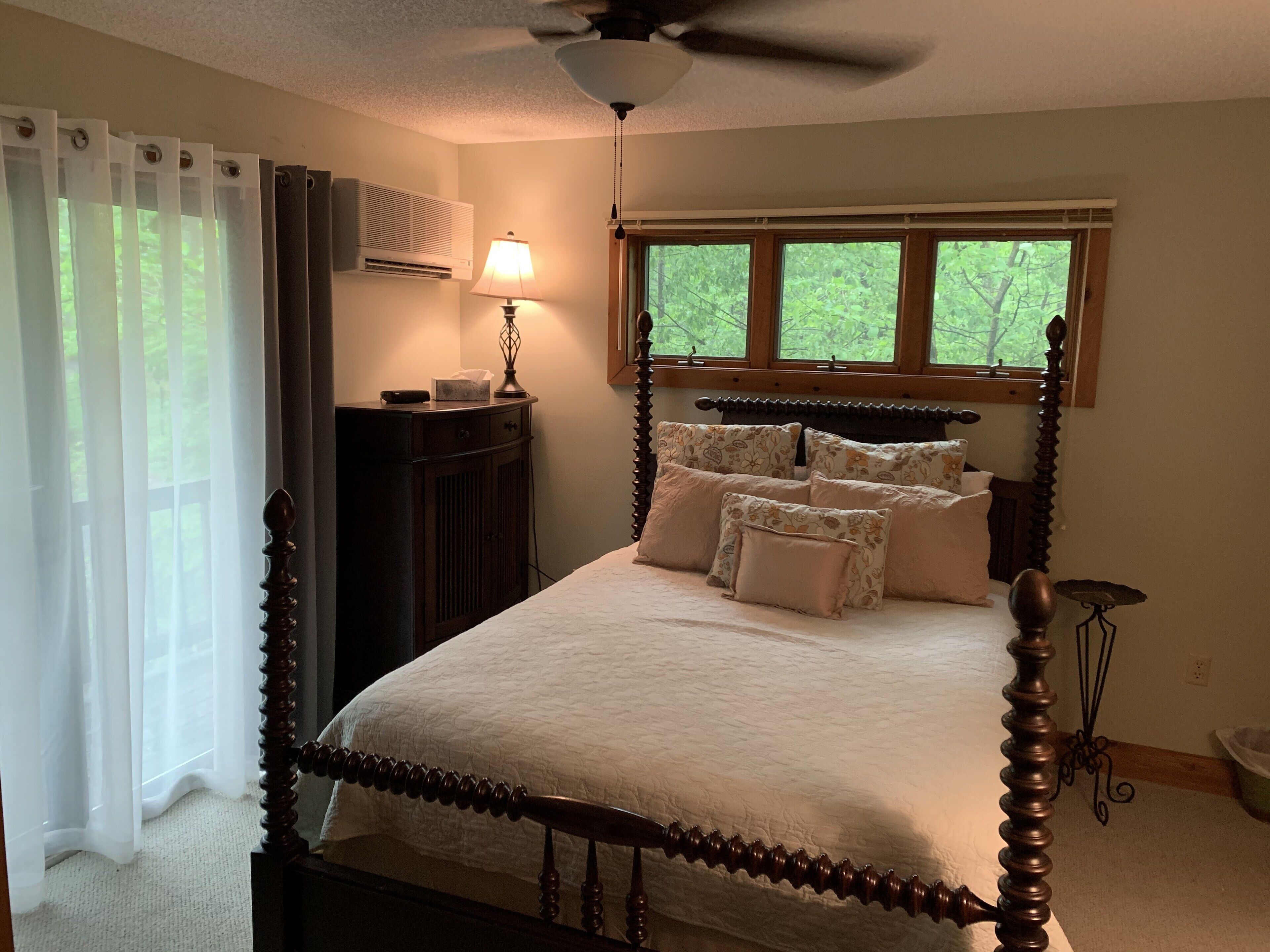 Quarto com cama de dossel, luminária de cabeceira, janela com vista para o verde e ventilador de teto.