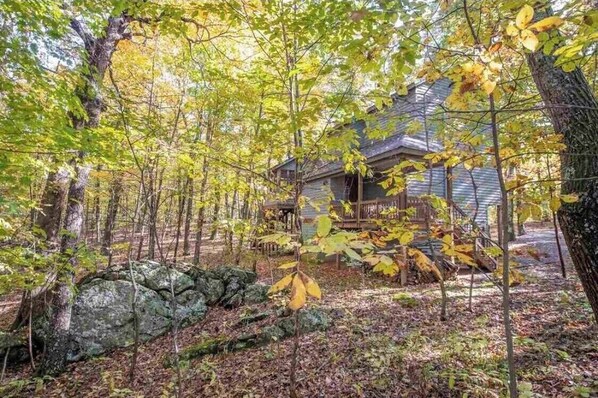 A rustic cabin surrounded by a forest with a stone wall and a wooden deck.