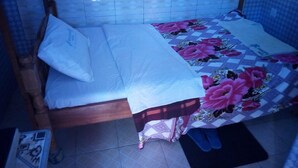 A small, single-bed room with a wooden frame, a white bedspread, and a floral-patterned blanket. The room has tiled walls and a tiled floor.