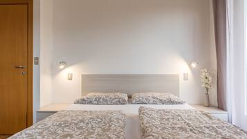 Ein Hotelzimmer mit Holztür, zwei Nachttischen, einem Bett mit gemusterten Betttextilien, einem Beistelltisch mit Blumenvase und einem Fenster mit Vorhängen.