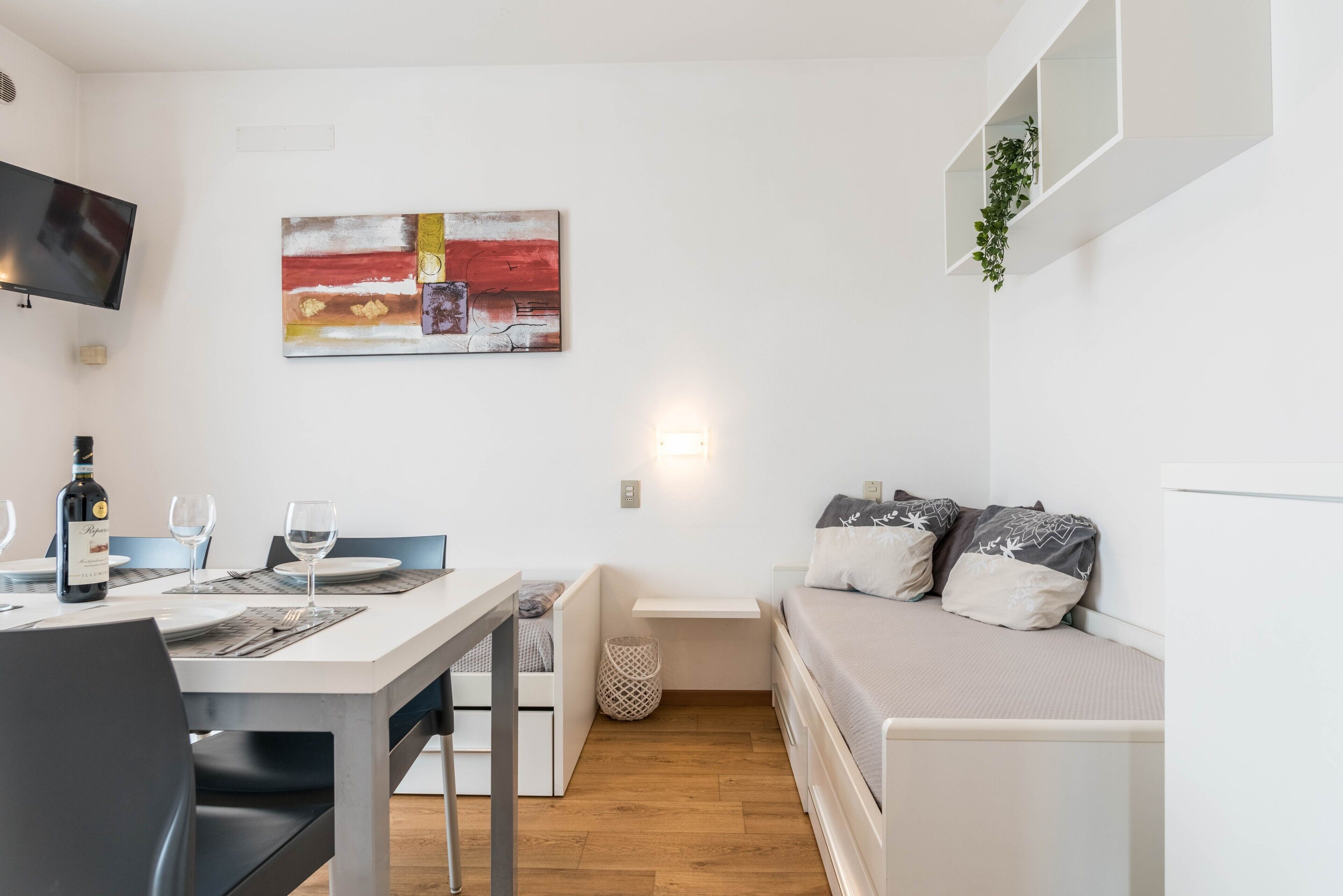 Un espace de vie compact comprenant un coin repas, un lit avec des oreillers, un téléviseur fixé au mur et une étagère murale agrémentée d’une plante.