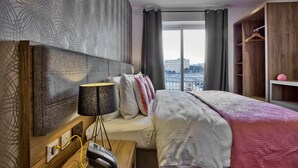 Habitación de hotel con una cama grande, una mesita de noche con lámpara, un armario de madera y vistas a la ciudad desde la ventana.