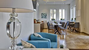 Una sala de estar moderna con un sofá azul, una mesa de centro de vidrio transparente y una lámpara. En el fondo, hay un área de comedor con una mesa y sillas.