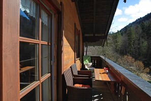 En stuga byggd av trä med stora fönster, en träveranda och utsikt över en skogsklädd dal.