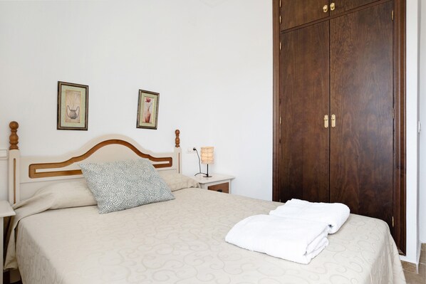 A neatly made bed with a patterned bedspread, a wooden headboard, two framed pictures on the wall, a bedside lamp, and a wooden wardrobe.