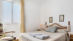 A neatly made bed with a patterned bedspread, a chair, a bedside table with a lamp, and a window with curtains.
