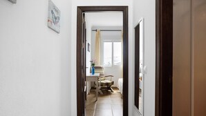 A hallway leading to a room with a bed, a chair, a table, and a window with curtains.