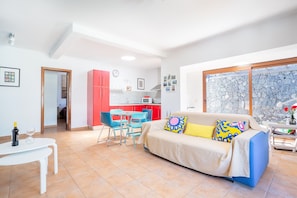 Ein Wohnzimmer mit einer Couch, einem Esstisch und Stühlen. Im Hintergrund befindet sich eine Küche mit roten Schränken und einer weißen Arbeitsplatte.