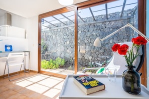 Ein Zimmer mit Schreibtisch, Stuhl, Buch, Blumen in einer Vase, Lampe und Blick auf eine Mauer aus Stein und grüne Bepflanzung draußen.