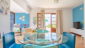 Een moderne woonkamer met een eettafel met glazen blad, blauwe stoelen, een bank, een televisie en uitzicht op de omgeving.