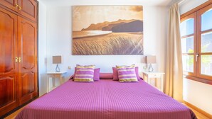 Een slaapkamer met een groot bed, een houten kledingkast, een schilderij van een kustlandschap en een raam met gordijnen.