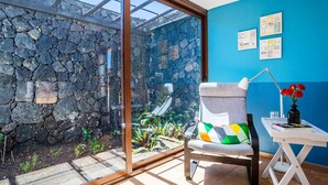 Een kamer met een stenen muur, een glazen deur naar een tuin, een stoel met een kleurrijk kussen, een kleine tafel met een lamp en een bloemenvaas, en een blauwe muur met ingelijste foto's.