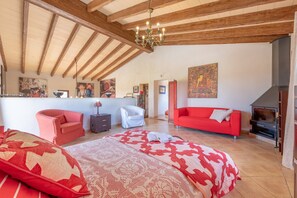 A spacious room with a high ceiling, wooden beams, a large bed, a red sofa, a white armchair, a fireplace, and artwork on the walls.