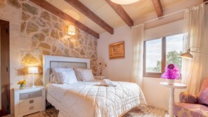 A bedroom with a stone wall, a bed with a white headboard, a nightstand with a lamp, a window with curtains, and a chair with a purple cushion. - Majorcan Finca with Pool - Son Draguet (Porreres)