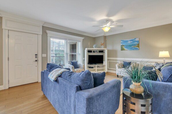 A living room with a blue sofa, a TV stand, and a ceiling fan. - Heated Pool Access | 5*Clean | Flex Cancelation (Tybee Island)