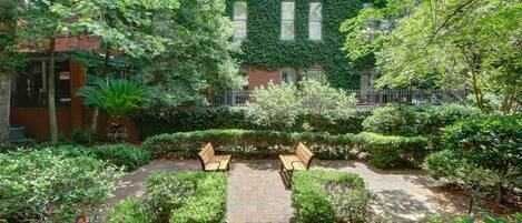 A garden courtyard with a paved walkway, two wooden benches, and a brick building with ivy-covered walls.