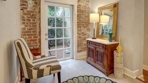 A room with a brick wall, a wooden cabinet, a striped chair, a door leading outside, and a mirror with a lamp. - Heated Pool Access | 5*Clean | Flex Cancelation (Savannah)