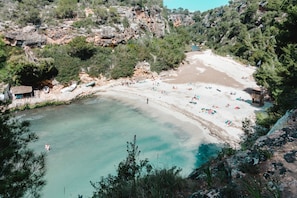 A coastal beach with clear water, rocky cliffs, and a sandy area with umbrellas.