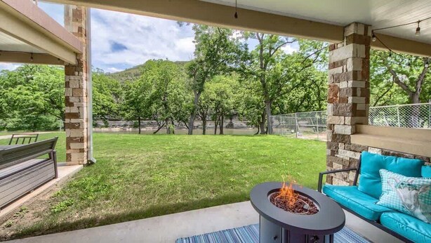 An outdoor patio area with a fire pit, a blue sofa, and a view of a grassy yard and trees. - RG 111 Guadalupe Fortress of Awesome (New Braunfels)