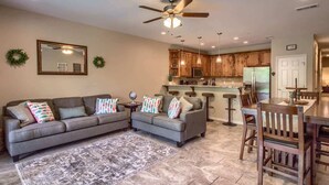 A living room with a sectional sofa, a coffee table, a dining table with chairs, a kitchen area with wooden cabinets, a ceiling fan, and a large mirror on the wall. - RG 111 Guadalupe Fortress of Awesome (New Braunfels)