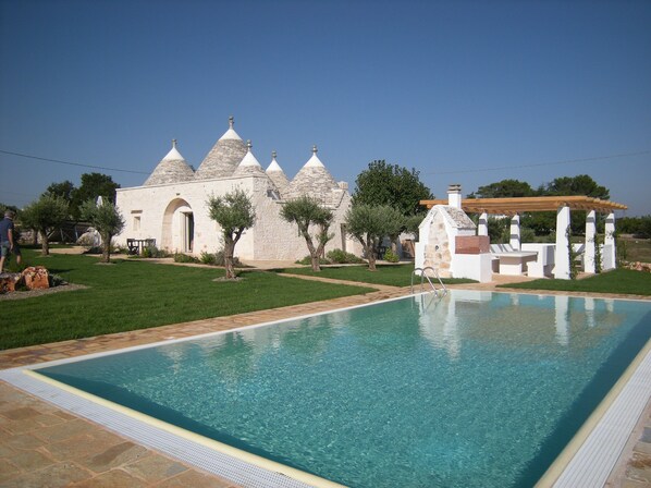 A swimming pool with a stone deck, a white building with multiple conical roofs, and a pergola with seating.