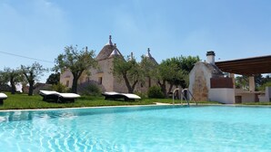 A swimming pool with lounge chairs, a stone building with multiple roofs, and trees in the background. - Luxury Villa "Trulli Terrarossa" in Apulian style with Pool (Martina Franca)
