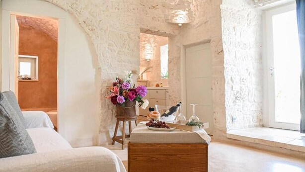 A rustic room with a stone wall, a wooden bench, a small table with a vase of flowers, and a bed with a pillow. - Luxury Villa "Trulli Terrarossa" in Apulian style with Pool (Martina Franca)
