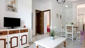 Ein Wohnzimmer mit Fernseher auf einer Kommode, ein Essbereich mit Tisch und Stühlen und eine Küche im Hintergrund.