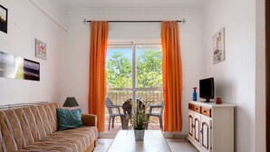Ein Zimmer mit einer Couch, einem Tisch, einem Schrank, einem Fernseher und einem Fenster mit Vorhängen.