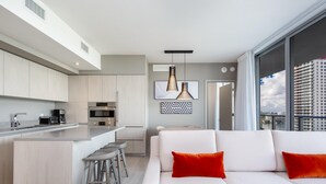 Cocina moderna con una isla central, taburetes de bar y una zona de comedor con sofá blanco y cojines rojos.
