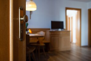 A wooden door with a lock, a kitchen area with a dining table and chairs, and a television on a stand. - Cosy Apartment 1 "Zur Sonne" with Mountain View & Garden (Lajen)