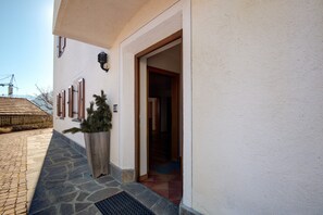 Un bâtiment dont l’entrée est pavée de pierres, doté de volets en bois et agrémenté d’un jardinier en bois où poussent des plantes vertes.
