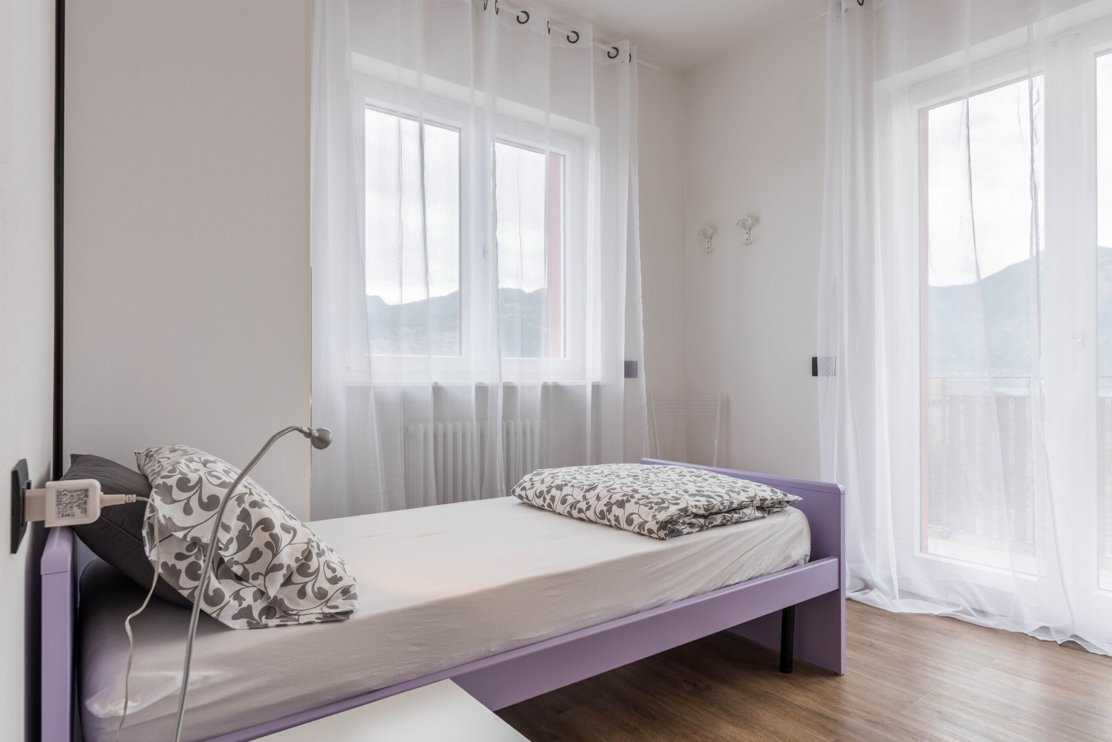 Ein Einzelbett mit Kopfteil, ein Nachttisch, ein Fenster mit transparenten Vorhängen und Blick ins Freie.