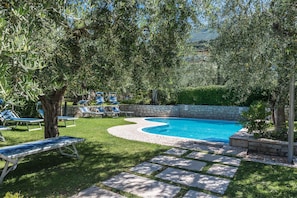 Une piscine bordée de pierres, entourée de pelouse et d’arbres, avec des chaises longues et un mur en pierre.