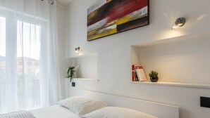 A modern hotel room with a large bed, a window with sheer curtains, a wall-mounted abstract painting, and a small shelf with a plant and books.