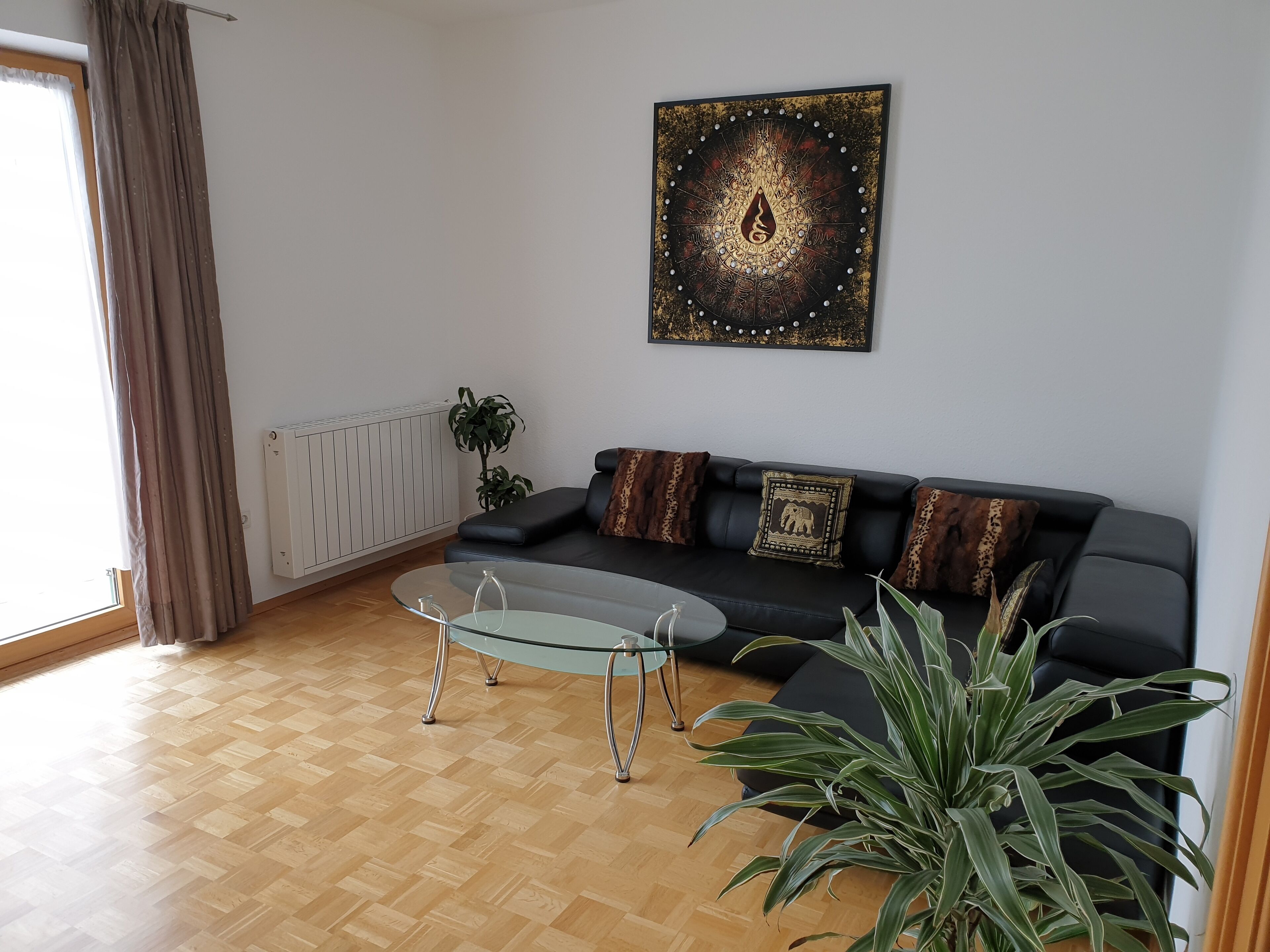A living room with a black leather sofa, a glass coffee table, a potted plant, and a wall-mounted artwork.