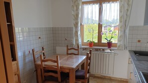 Ett kök med kakel på väggen bakom arbetsbänken, ett träbord och stolar, ett fönster med krukväxter och en inbyggd ugn.
