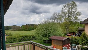 En balkong med utsikt över en grön äng, ett trähus och en röd bil som står parkerad längre bort.