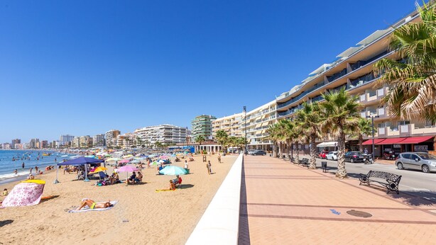 Um calçadão à beira-mar com uma praia de areia, guarda-sóis e uma fileira de edifícios com varandas.