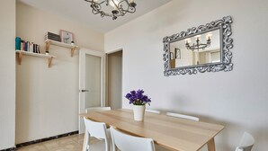 Uma sala de jantar com uma mesa de madeira, cadeiras brancas, um vaso com flores roxas, um espelho com moldura elaborada e estantes com livros e objetos decorativos.