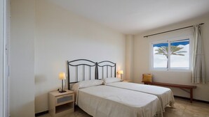 Quarto com duas camas, uma mesinha de cabeceira, um banco, janela com vista e cortina.