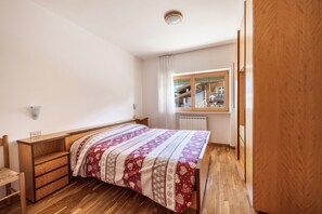A bedroom with a wooden bed, a desk, a chair, a radiator, and a window with a view of a building.