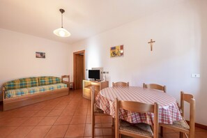 A room with a sofa, a dining table with chairs, a television on a stand, and a cross on the wall.
