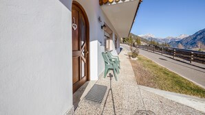 Un edificio con facciata bianca, una porta in legno con maniglia a forma di cuore, un balcone con ringhiera di legno e una vista chiara sulle montagne sullo sfondo.