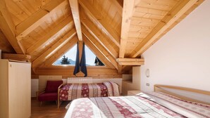 Un dormitorio con techo de madera inclinado, una cama con ropa de cama estampada, una silla roja y una ventana con vistas a árboles.