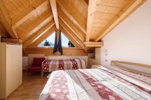 A bedroom with a sloped wooden ceiling, a bed with patterned bedding, a red chair, and a window with a view of trees.