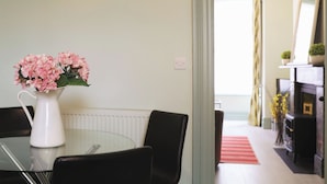 A dining area with a table, chairs, and a vase of pink flowers. There is a radiator on the wall and a window with curtains.